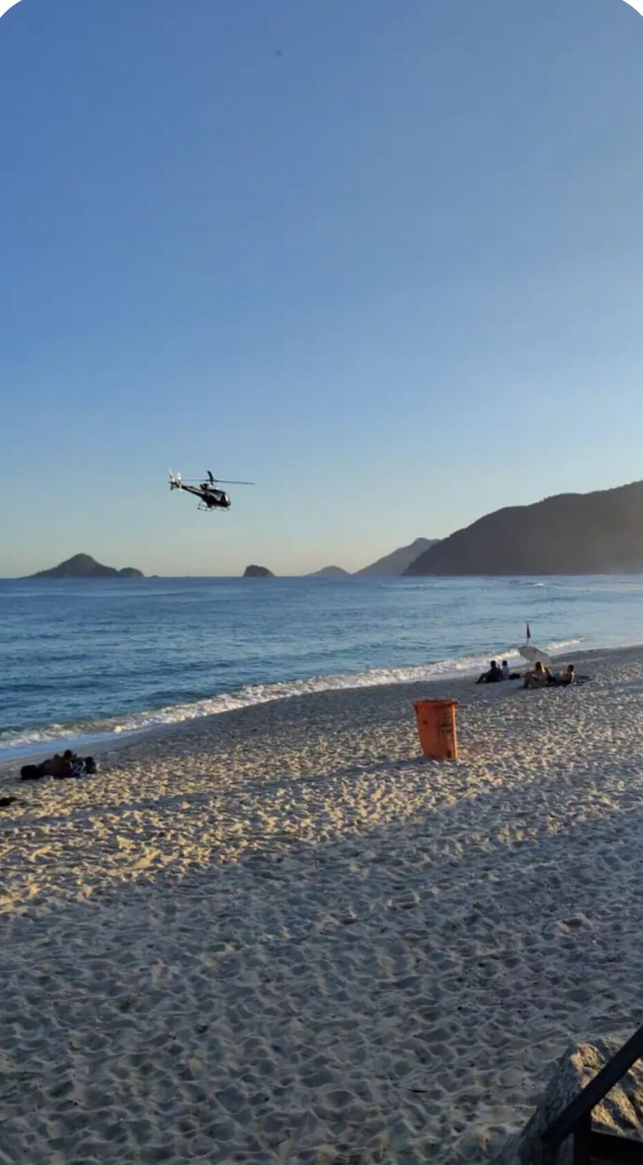 Ein Strand mit einem Hubschrauber über dem Kopf, ein paar Personen im Sand und einer Mülltonne.