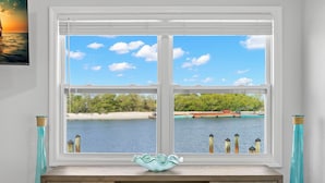 A room with a large window overlooking a beach and water, a wooden desk with a decorative bowl, and teal-colored vases on either side. - Turtle Nest (Sarasota)