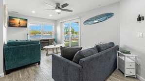 A modern living room with a gray sofa, teal armchair, and a white side table with a turtle figurine. A ceiling fan and a painting of a sailboat are visible. - Turtle Nest (Sarasota)