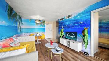 Een woonkamer met een strandmuurschildering, een televisie, een bank, een eettafel en een salontafel.