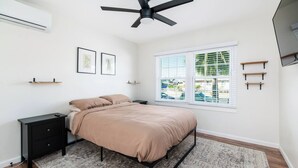Un dormitorio con cama, mesita de noche, ventilador de techo, ventana con persianas y vistas al exterior.
