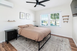 A bedroom with a bed, a nightstand, a ceiling fan, a window with blinds, and a view of the outside.