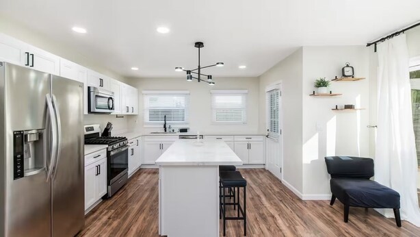 Una cocina moderna con armarios blancos, electrodomésticos de acero inoxidable, una isla central con taburetes y una silla negra cerca de la ventana.
