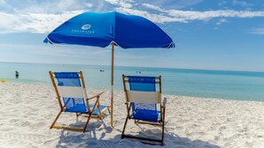 Twee strandstoelen en een blauwe parasol op een zandstrand met helderblauw water en enkele mensen in de verte.