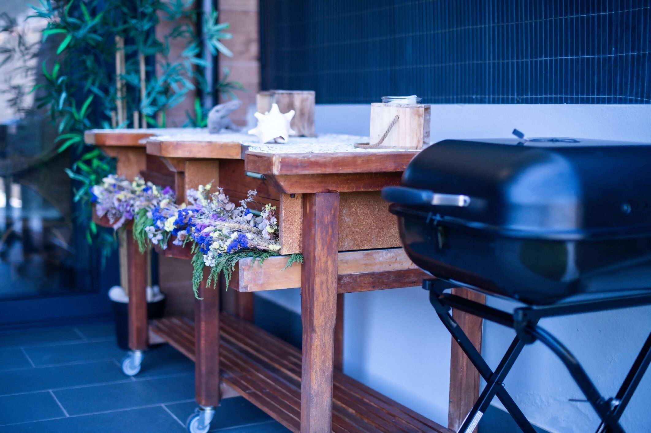 A wooden outdoor grill setup with a barbecue on wheels, positioned on a tiled patio with a potted plant and decorative items in the background.