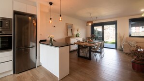 A modern kitchen with a stainless steel refrigerator, a dining area with a wooden table and chairs, and a large window providing a view of the outside. - Ribadesella House with Garden 2 km from Beach – Saelia (Ribadesella)