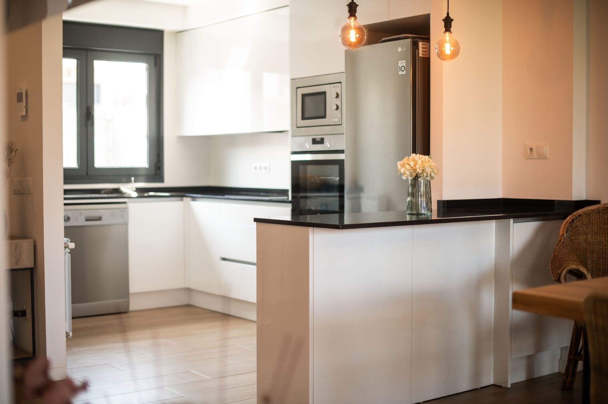 Une cuisine moderne avec des meubles blancs, un plan de travail noir, un four encastré, un four à micro-ondes et un réfrigérateur. On peut voir un vase rempli de fleurs sur le plan de travail et un luminaire suspendu au-dessus.