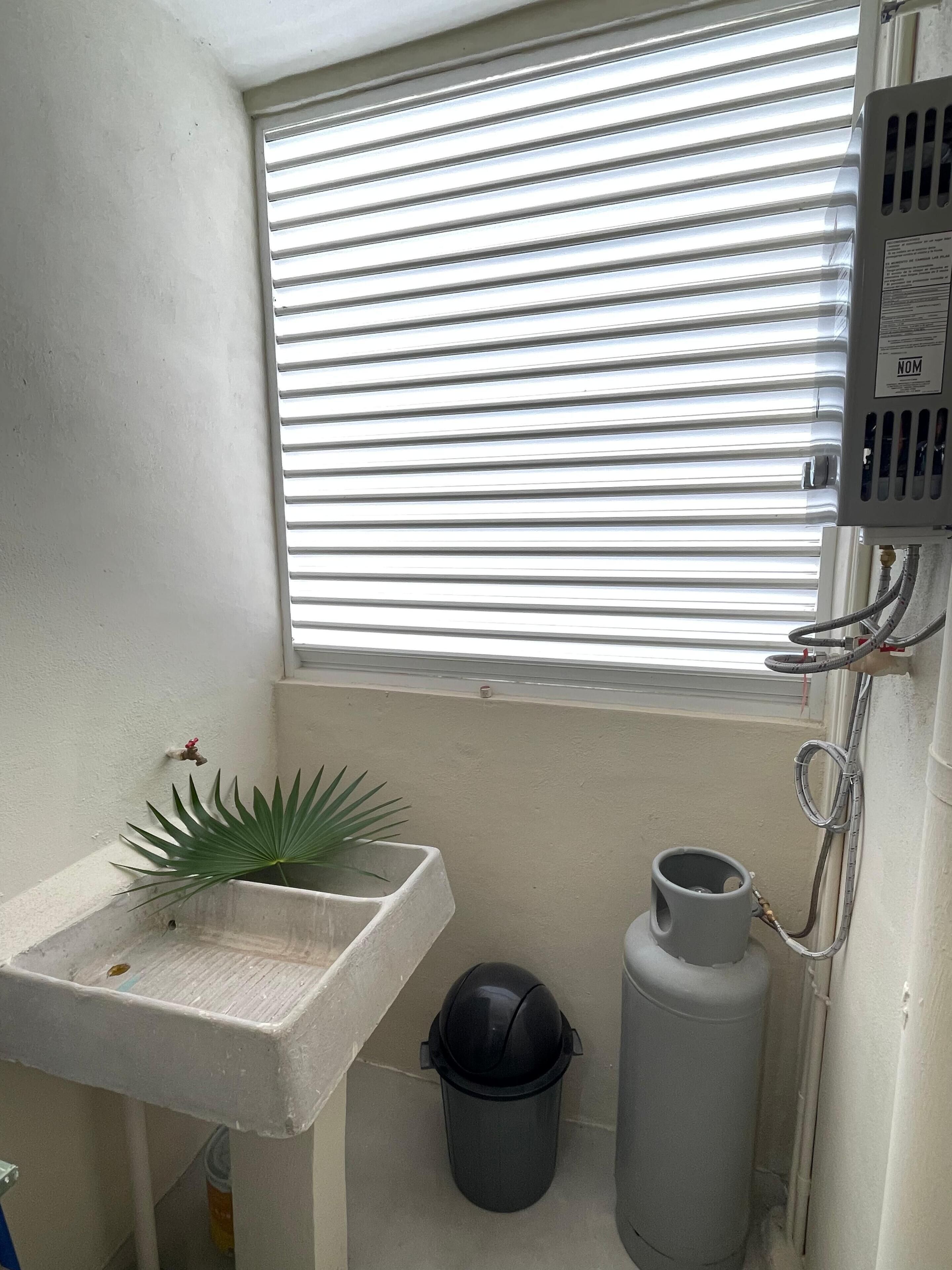 Ein kleines Badezimmer mit Fenster, Waschbecken, Mülleimer und Warmwasserbereiter.