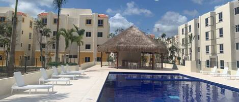 A resort pool area with lounge chairs, a thatched-roof structure, and multi-story buildings in the background.