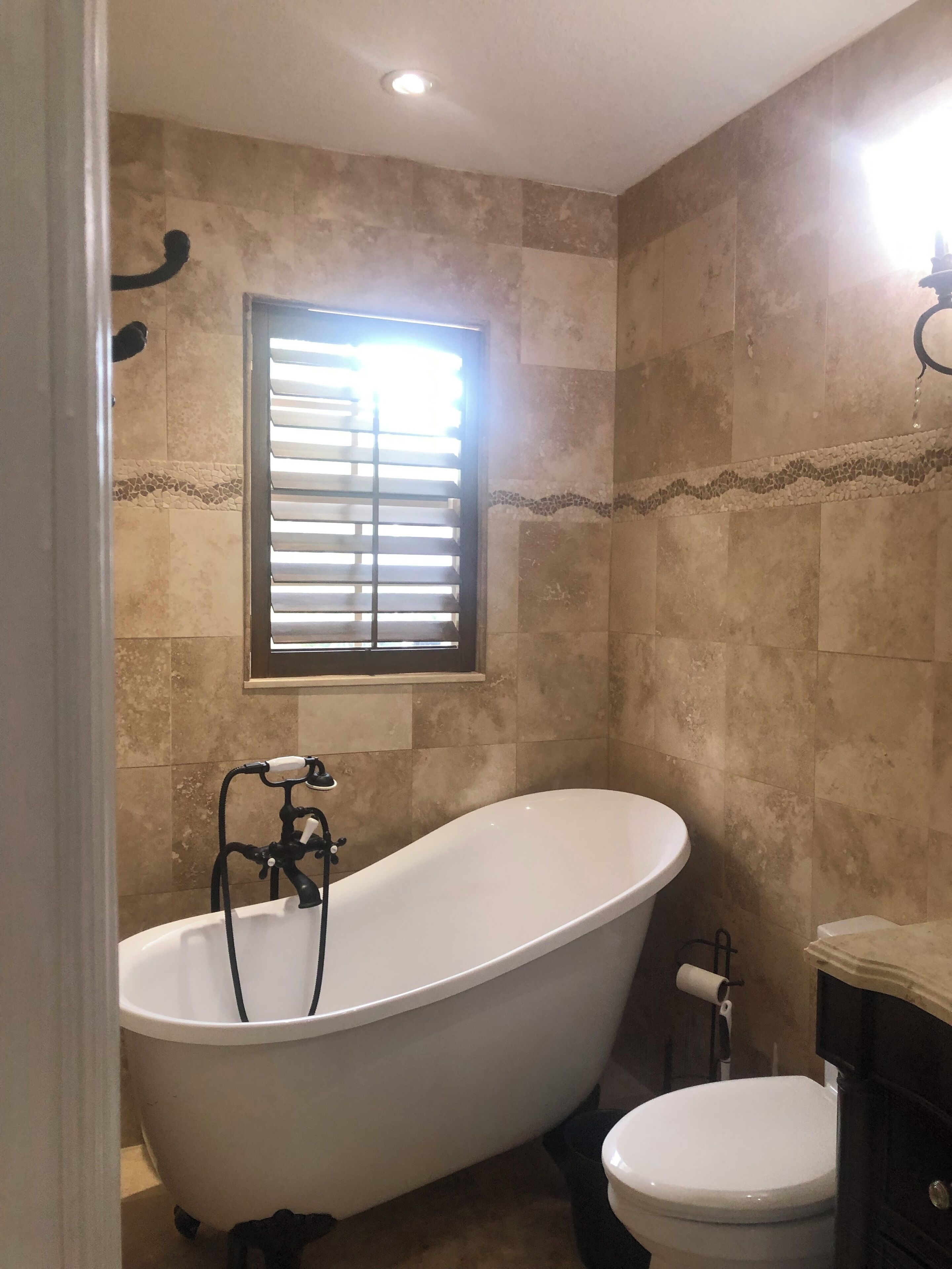 Een badkamer met een vrijstaand bad, toilet en een raam met zonwering.