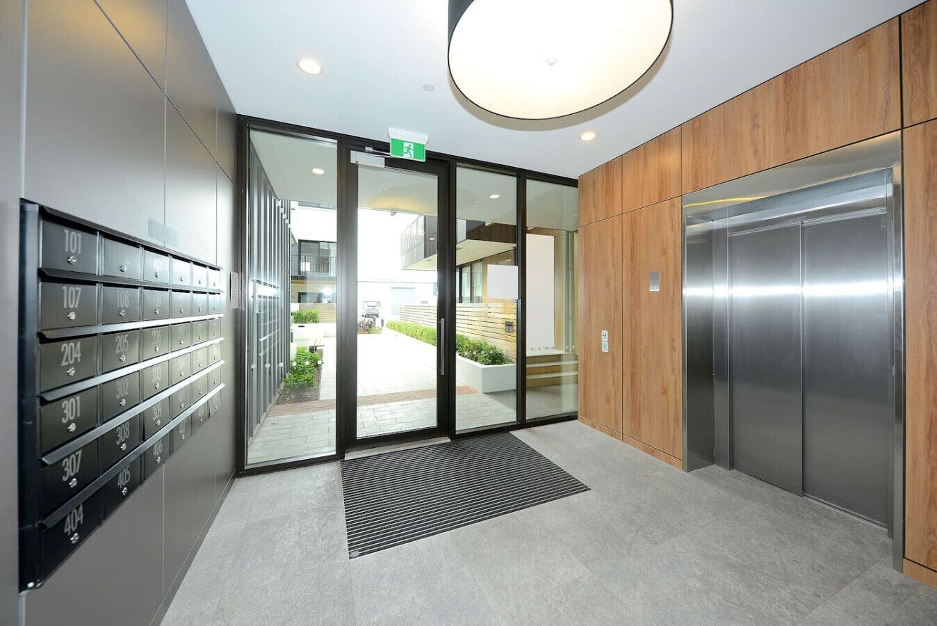 A modern elevator with a metallic finish, a glass door entrance with a green exit sign, and a series of mailboxes on the left.