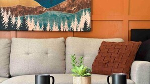 Een moderne woonkamer met een bank, twee zwarte koffiekopjes op een houten tafel, een kleine plant en een gebreid plaids kussen.