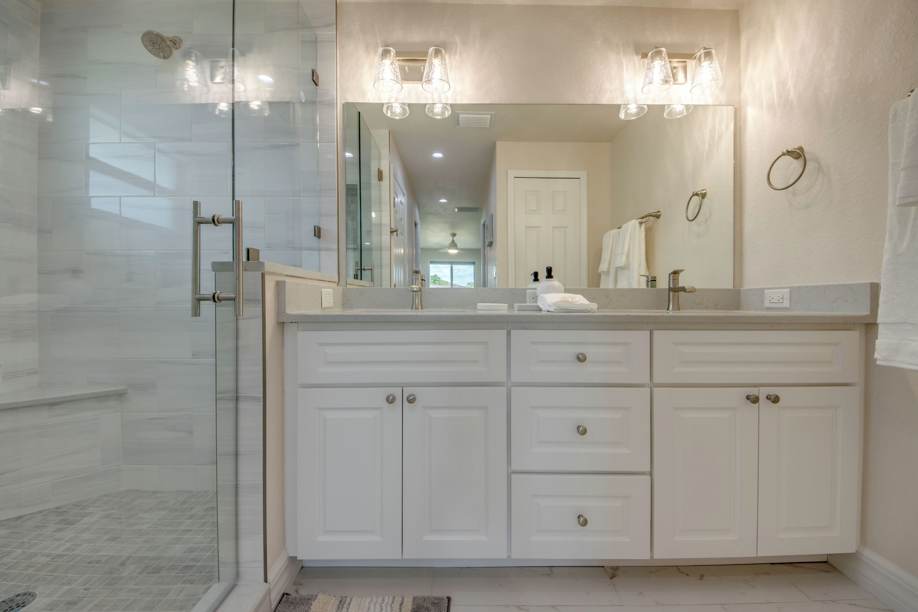 A modern bathroom with a glass shower enclosure, a large mirror with lights, and a white vanity with multiple drawers.
