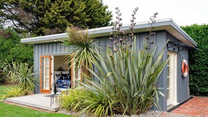 Une petite maison moderne avec une porte donnant sur un porche, entourée de différentes plantes et bordée d’un chemin en briques.