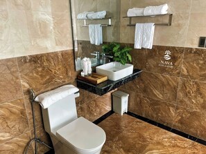 Ein Badezimmer mit Toilette, Waschbecken mit Spiegel darüber und einem Handtuchhalter mit Handtüchern. Auf der Ablage steht eine Pflanze und an der Wand das Logo von NAVA HOTEL & RESORT.