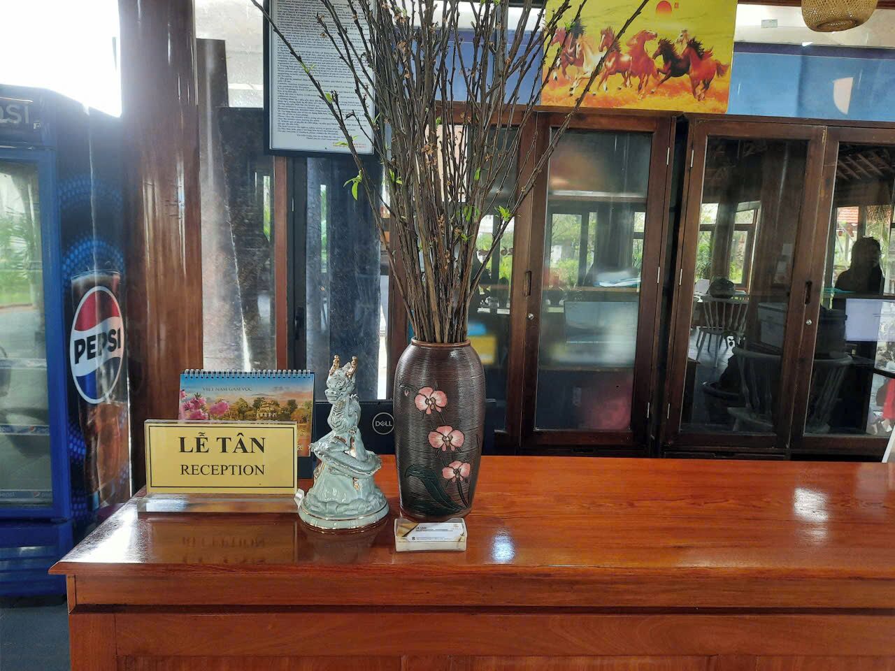 Un comptoir d’accueil avec une pancarte indiquant « Réception Lê Tân », un vase décoratif orné de fleurs roses et une petite statue.