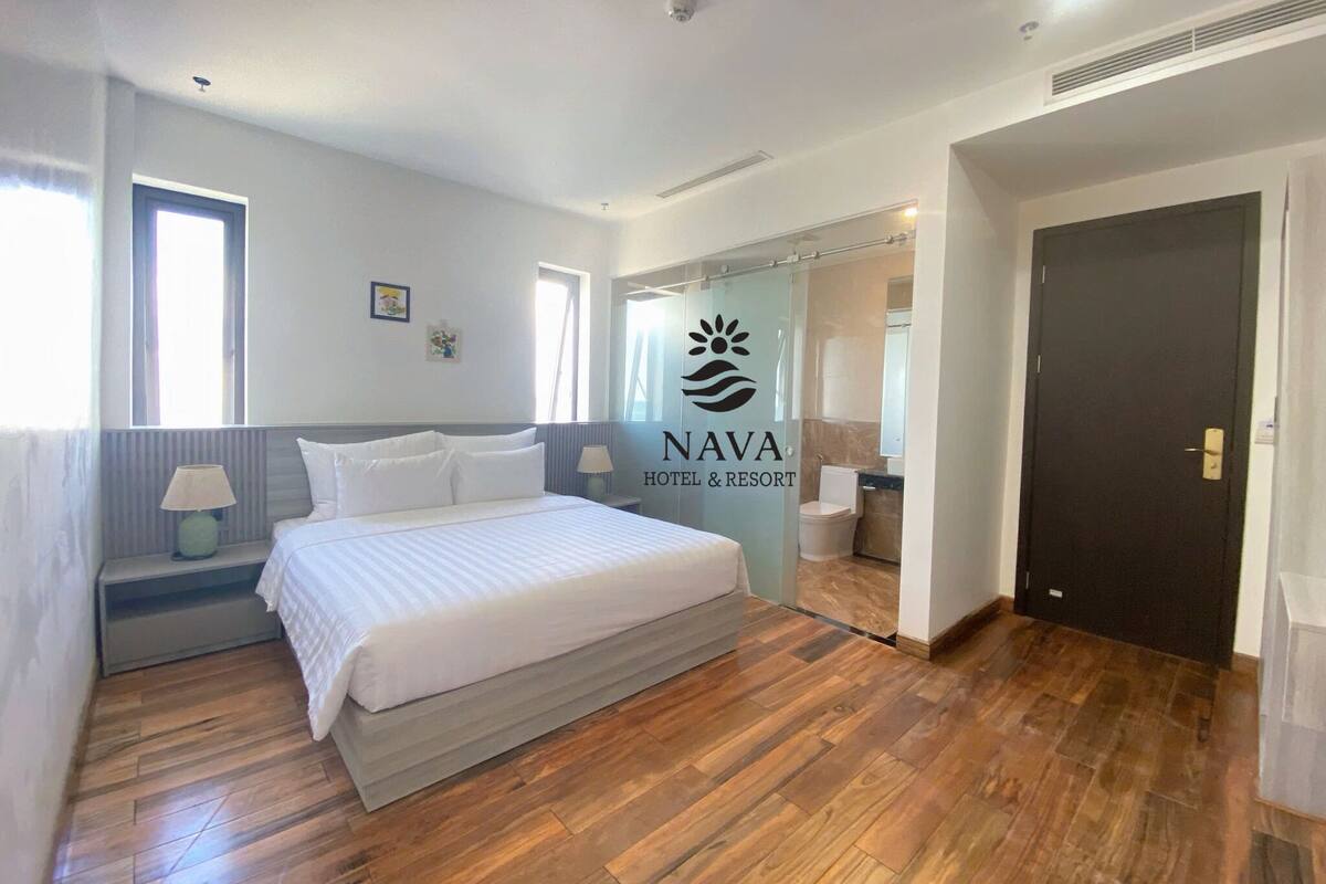 A hotel room with a large bed, a nightstand with a lamp, a window, and a bathroom visible through a glass partition.