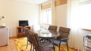 Ein modernes Wohnzimmer mit einem runden Glastisch, vier Stühlen, einem Flachbildfernseher, einem kleinen Bücherregal und einem Fenster mit Vorhängen.