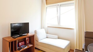 Ein kleines Wohnzimmer mit Fernseher, einem weißen Sofa, einem Holztisch und einem Fenster mit Vorhängen.