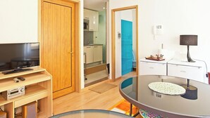 A compact hotel room with a wooden door, a television, a small desk with a computer, a dining table, and a kitchenette in the background.