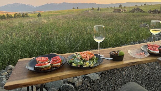 Una mesa de madera con platos de comida y copas de vino, con un fondo de campo cubierto de hierba y montañas al lejano.