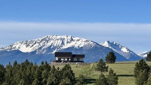 Una casa moderna con techo plano y ventanas grandes, rodeada por un campo cubierto de hierba y árboles, con montañas nevadas al fondo.
