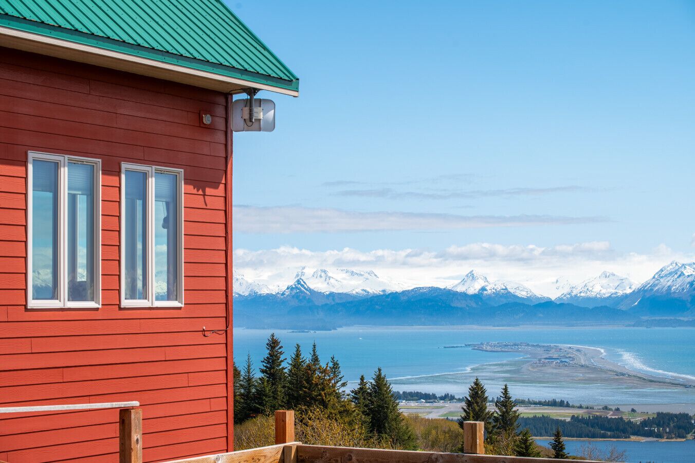 A red building with two windows and a green roof overlooking a coastal landscape with mountains and a body of water.