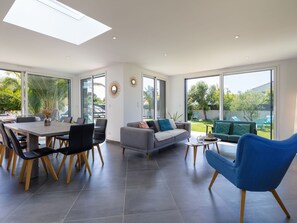 A spacious living area with a dining table, sofas, and chairs. Large windows provide views of the outdoors.