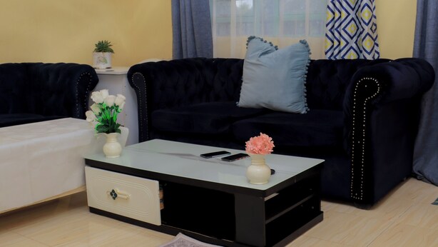 Ein Wohnzimmer mit einem dunkelgrauen Sofa, einem weißen Hocker, einem Glastisch mit einer Vase und einem Smartphone sowie einem kleinen Beistelltisch mit einer Topfpflanze.