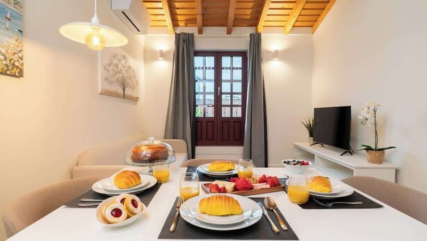 Une table blanche est dressée pour le petit-déjeuner, avec des croissants, des fruits et du jus. La pièce est dotée de poutres en bois, d’un téléviseur et d’une fenêtre agrémentée de rideaux.