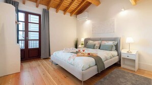 Une chambre avec un plafond en bois, un lit recouvert d’une literie à motifs floraux, une table de chevet avec une lampe et une fenêtre avec des rideaux.