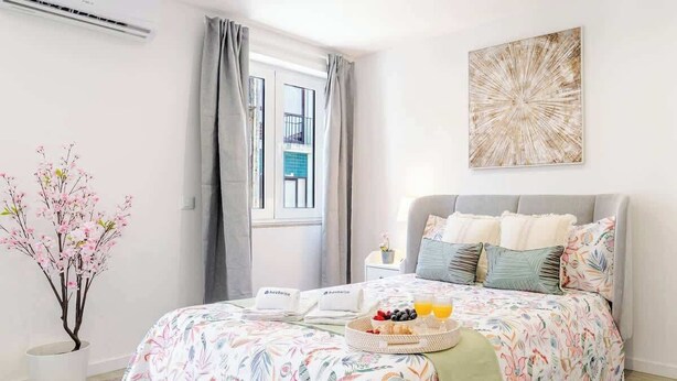 A bedroom with a floral bedspread, a bedside table with a tray of fruit and juice, a potted plant, and a view of the outside balcony. - Modern & Bright Studio by HostWise (Porto)
