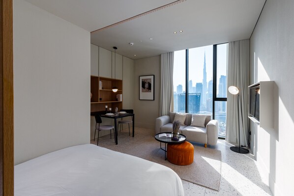 A modern hotel room with a large bed, a desk with a chair, a sofa, and a view of the city skyline.