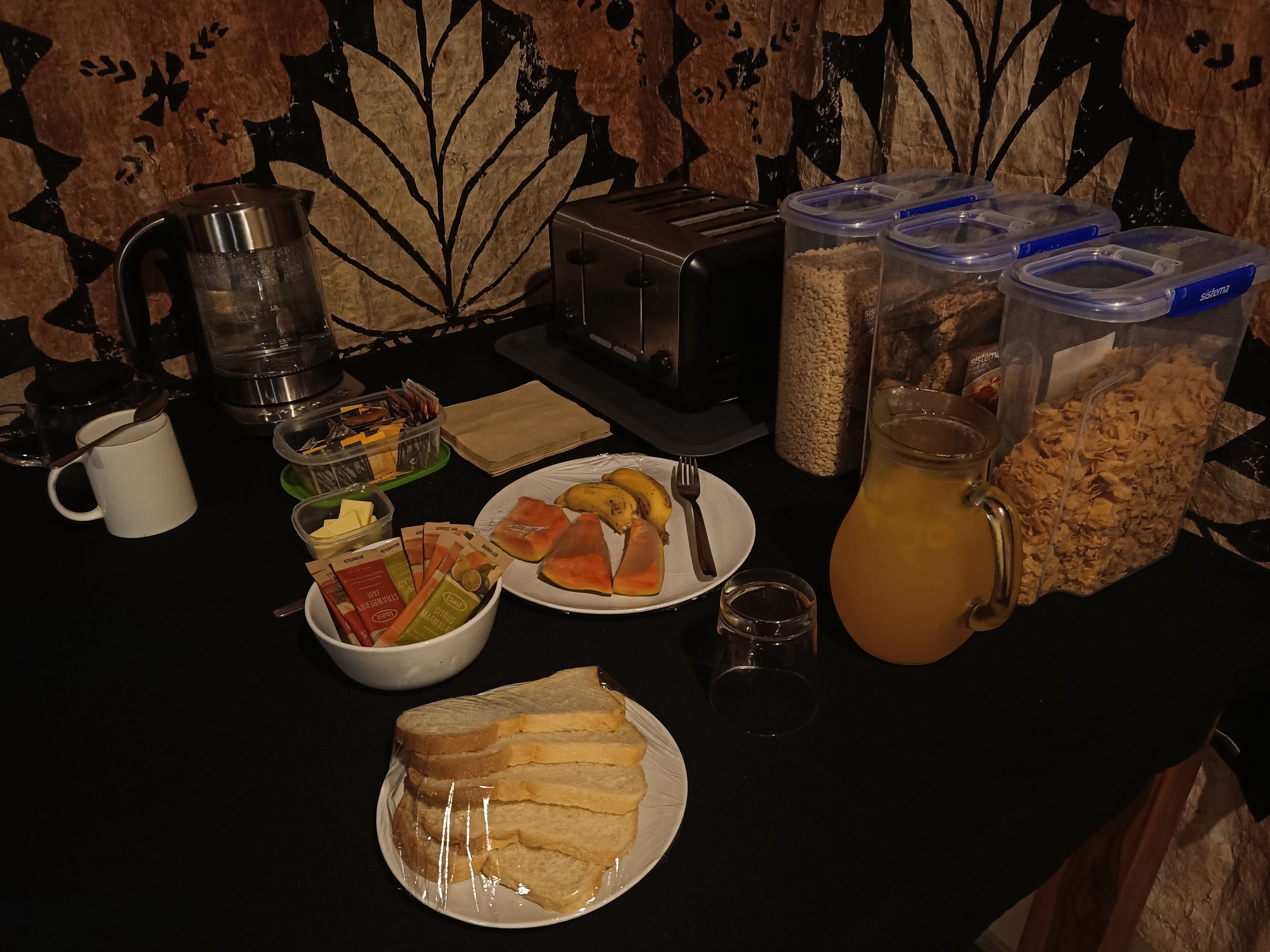 Una mesa de desayuno con cafetera, tostadora, recipientes de cereal, jarra de jugo de naranja, plato con tostadas y fruta, y un bol de cereal.