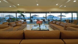 Un vaste espace salon avec plusieurs canapés disposés autour d’une table basse centrale, de grandes fenêtres offrant une vue sur la mer et les montagnes, et des parasols à l’extérieur pour créer de l’ombre.