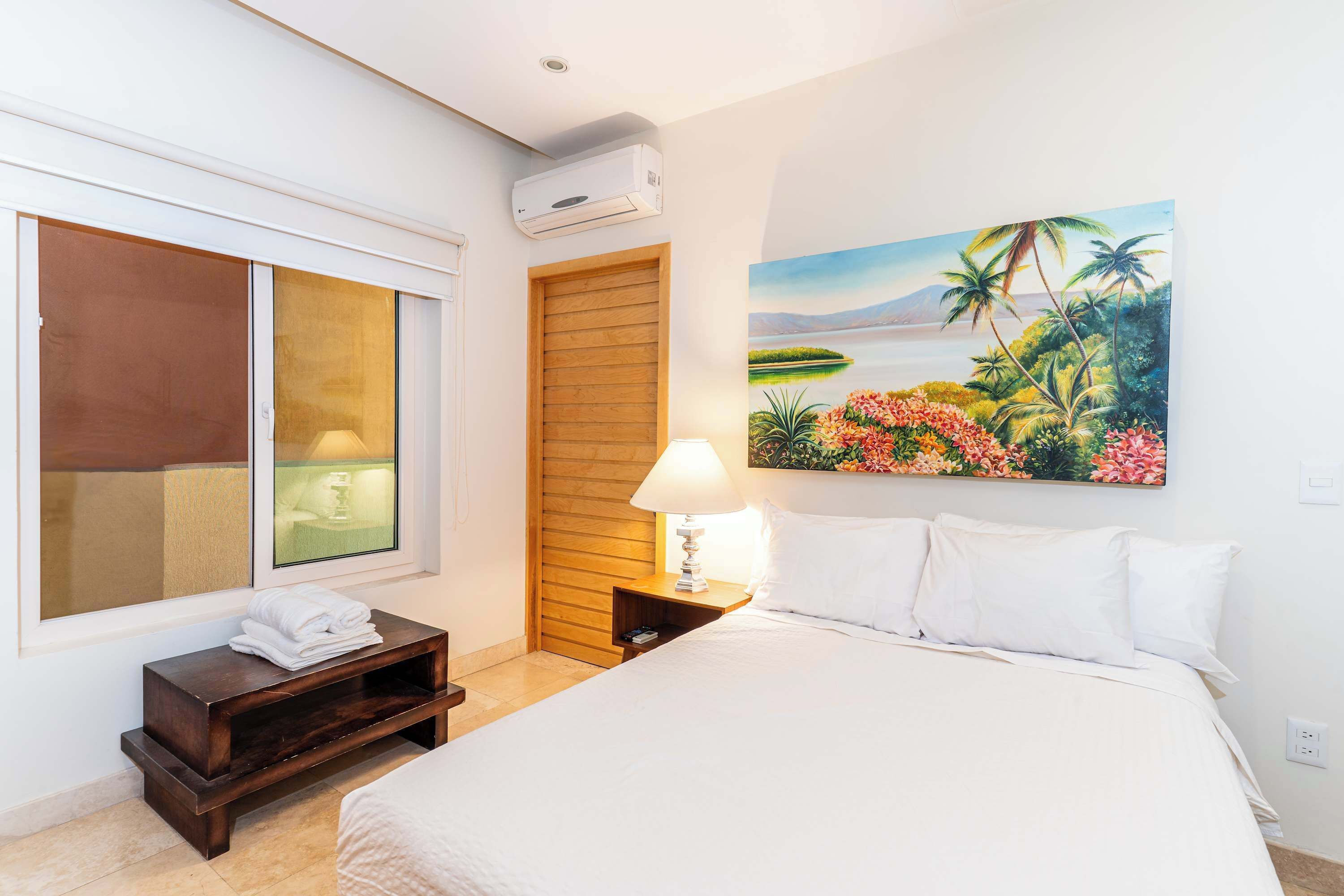 A hotel room with a bed, a nightstand with a lamp, a window with blinds, a painting of a tropical landscape, and a wooden door.