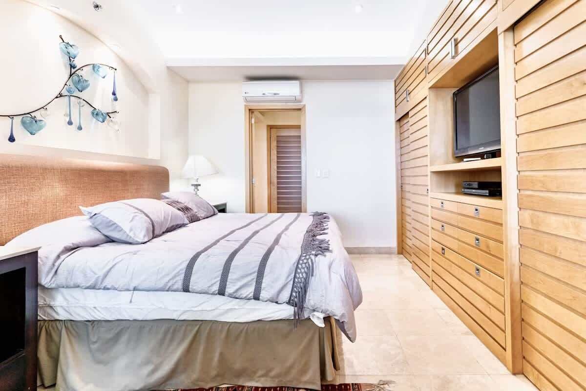 A bedroom with a bed, a TV on a wooden cabinet, and a wall-mounted air conditioning unit.