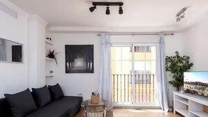 A modern living room with a black sofa, a small round table, a large window with curtains, and a TV on a stand.