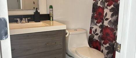 Un baño con inodoro, lavamanos con espejo, un mueble sobre el lavamanos y cortina de ducha con estampado floral.