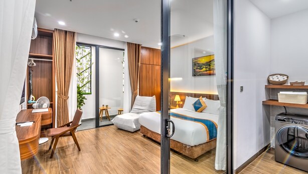 Ein modernes Hotelzimmer mit Holzboden, einem Bett mit blau-weißer Bettdecke, einem Schreibtisch mit Stuhl, einem kleinen Tisch und einer Waschmaschine.