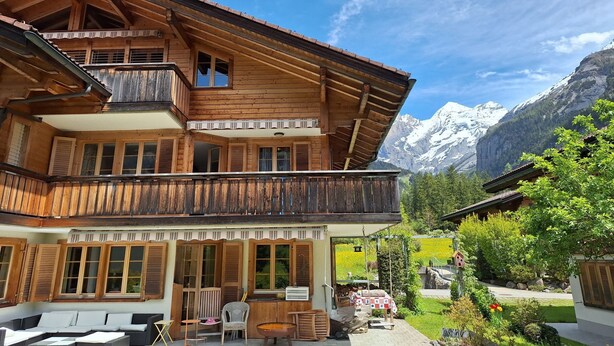 A wooden chalet with a balcony, outdoor seating, and a view of snow-capped mountains. - Haus Europa by Interhome (Kandersteg)