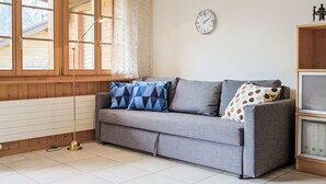 A room with a grey sofa, patterned cushions, a wall clock, and a radiator. - Haus Europa by Interhome (Kandersteg)