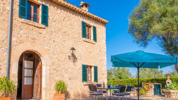 Un bâtiment en pierre doté d’une porte et de fenêtres, un espace extérieur aménagé avec des chaises et une table, et un grand parasol.