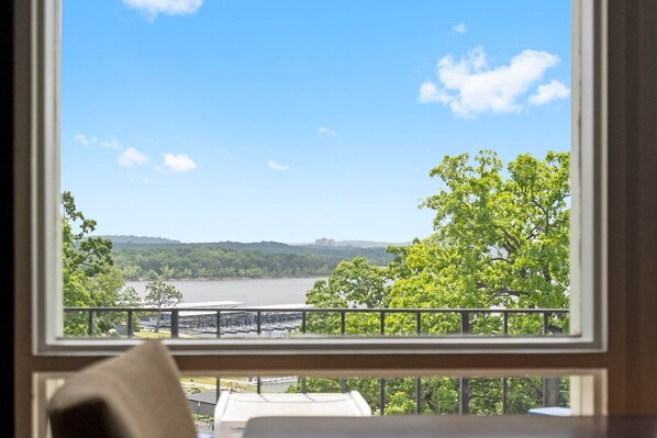 A room with a view of a lake, trees, and a distant building.