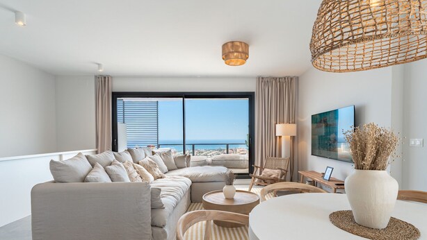 Un salon moderne doté d’un grand canapé d’angle, d’une table basse ronde, d’un luminaire suspendu en osier et offrant une vue sur la mer grâce à de grandes fenêtres du sol au plafond.