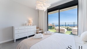 Une chambre moderne avec un grand lit, une commode blanche, une lampe de chevet et une vue sur la mer par une grande fenêtre.