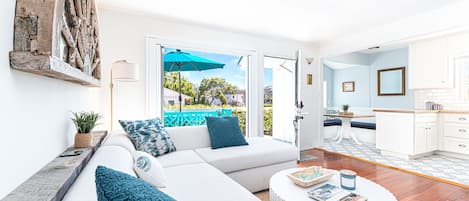 Ein geräumiges Wohnzimmer mit einem weißen Sofa, blauen Kissen, einem runden Couchtisch und Blick ins Freie.