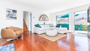 Una sala de estar moderna con un sillón de cuero marrón, un sofá blanco con cojines azules y una mesa de centro redonda. En la pared, hay un cuadro enmarcado, un espejo con marco de madera y una escalera que conduce a otra habitación.