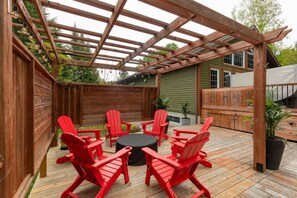 A wooden deck with a pergola, red Adirondack chairs, and a round table.