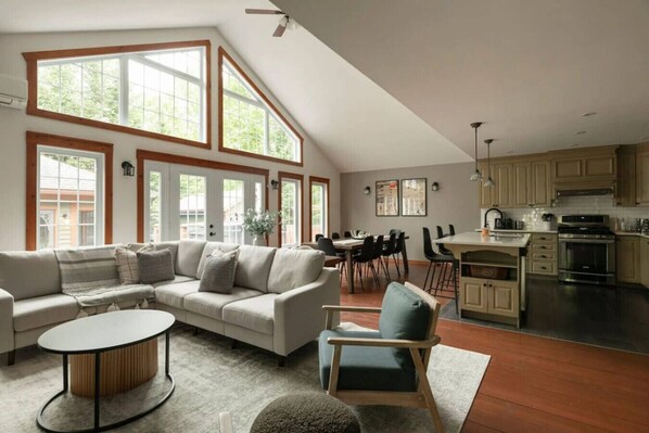 A spacious living area with a high ceiling, large windows, a dining table, and a kitchen area.
